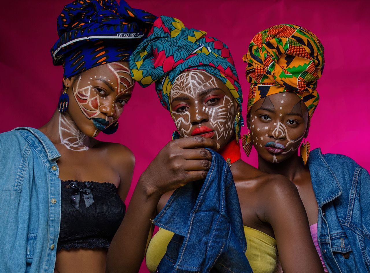 Home Striking portrait of three women showcasing African fashion with colorful headwraps and intricate face paint.