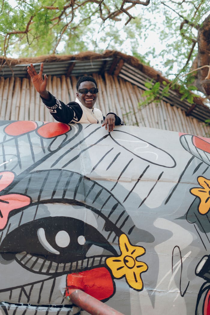 Home Young man joyfully waves from behind a vibrant mural depicting expressive art outdoors.