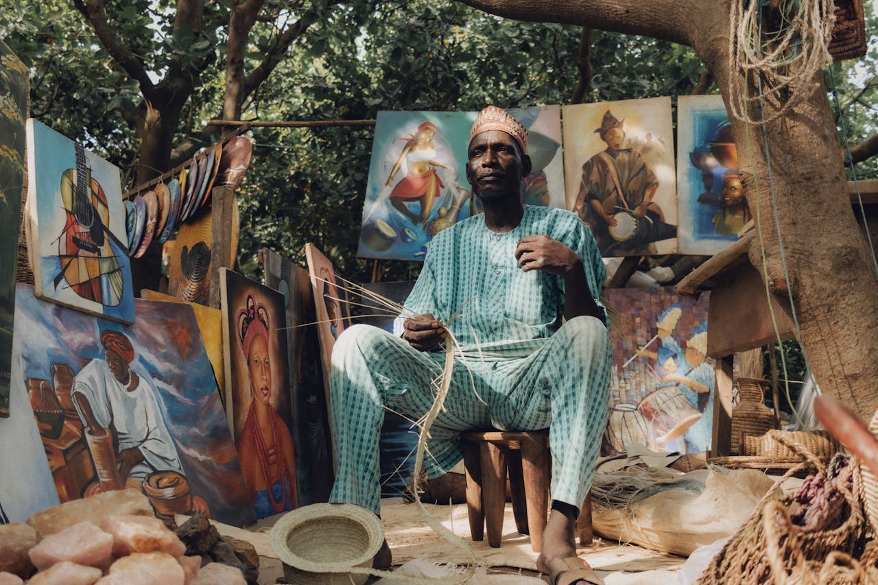 hero-img Explore the vibrant Nigerian art and craft market with traditional paintings and a skilled artisan at work.