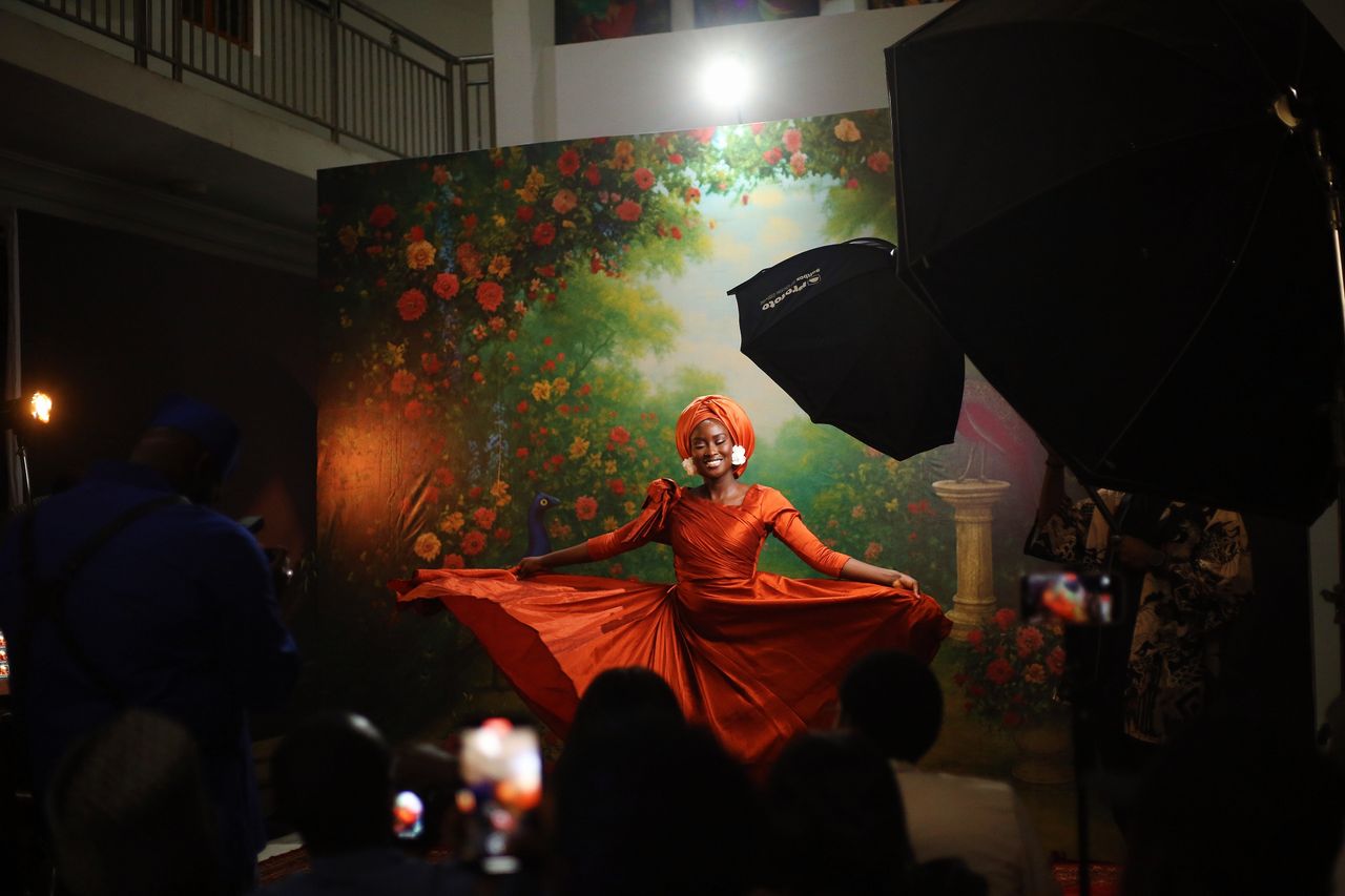 Home A woman in an orange dress poses gracefully indoors with a vibrant floral backdrop in Abuja, Nigeria.